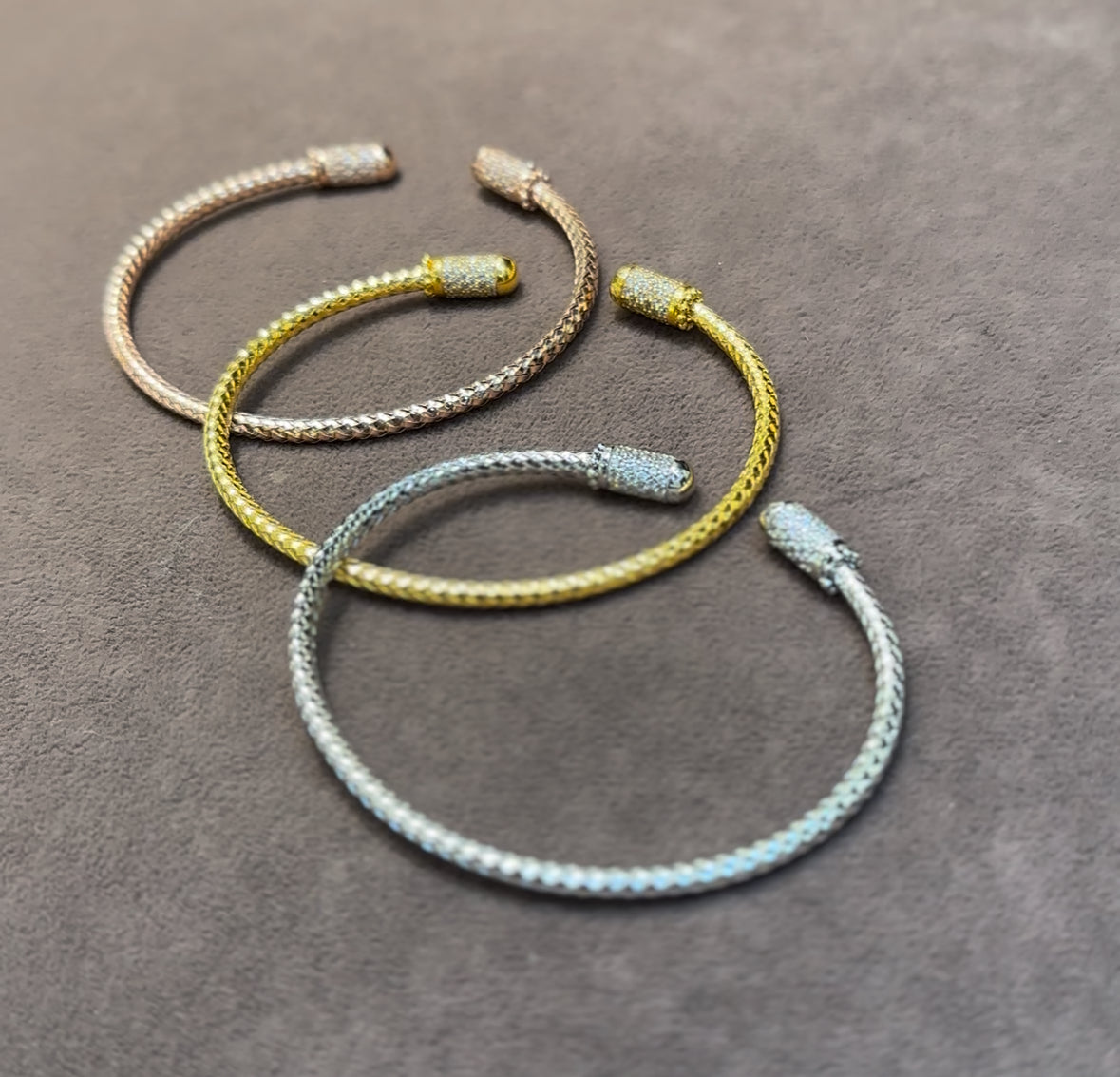 Three braided bracelets in gold, silver, and bronze on a gray surface
